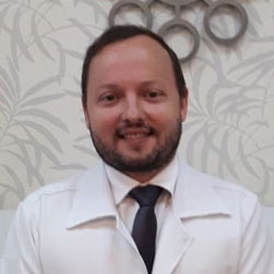 médico sorri para camera de jalelco branco e gravata preta com barba e logotipo da clinica ao fundo