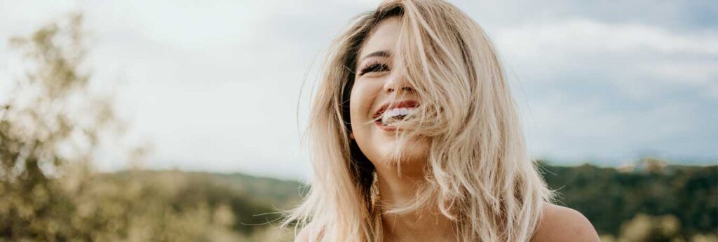 mulher loira cerca de 40 anos sorri com cabelo no rosto em meio a natureza