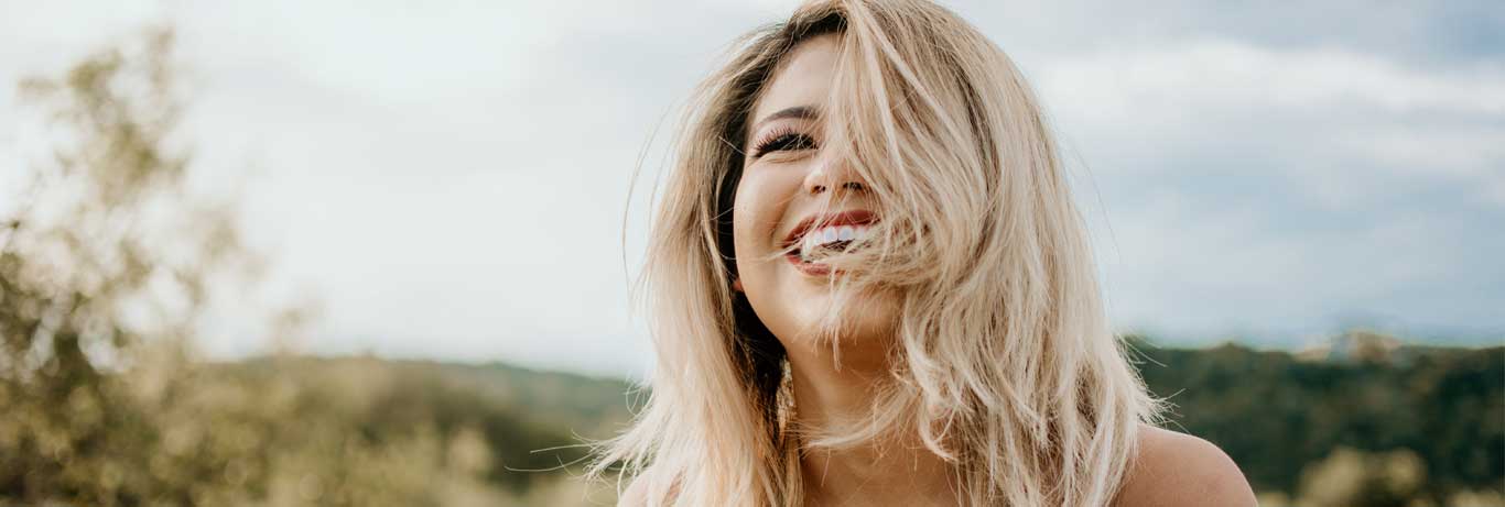 mulher loira cerca de 40 anos sorri com cabelo no rosto em meio a natureza