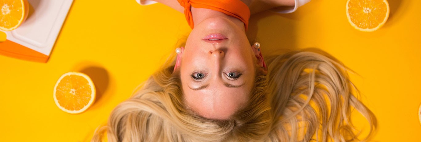 mulher loira deitada de cabeça pra baixo sobre fundo laranja com algumas laranjas ao redor