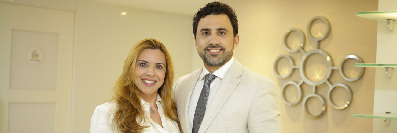 mulher de cabelo loiro na altura do peito sorrindo de branco ao lado de homem com barba sorrindo de terno claro com logo da leger ao fundo