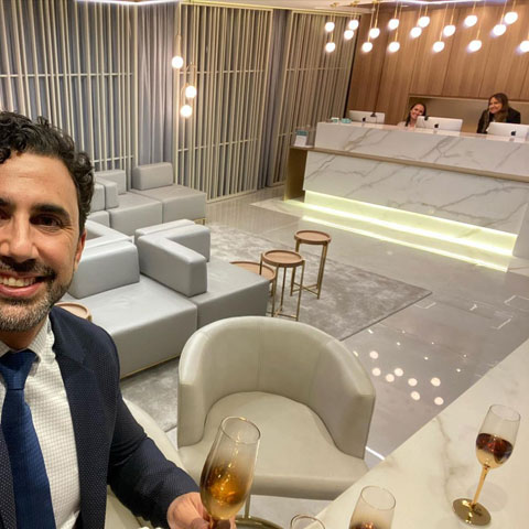 doutor roberto chacur com taça de champagne na mão tira selfie na recepção com atendentes sentadas ao fundo sofá central e poltronas ao redor da sala em tons de branco e dourado