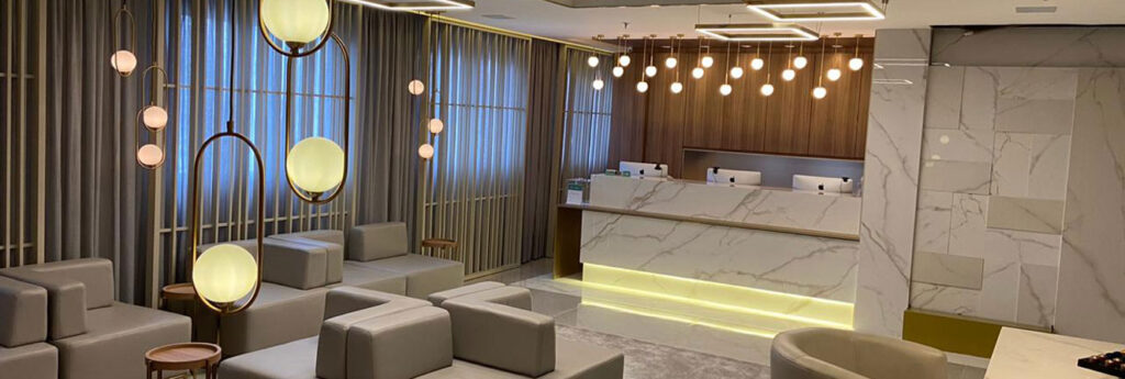 sala de recepção com sofás no centro e poltronas ao redor com balcão de recepção ao fundo em tons brancos e dourados