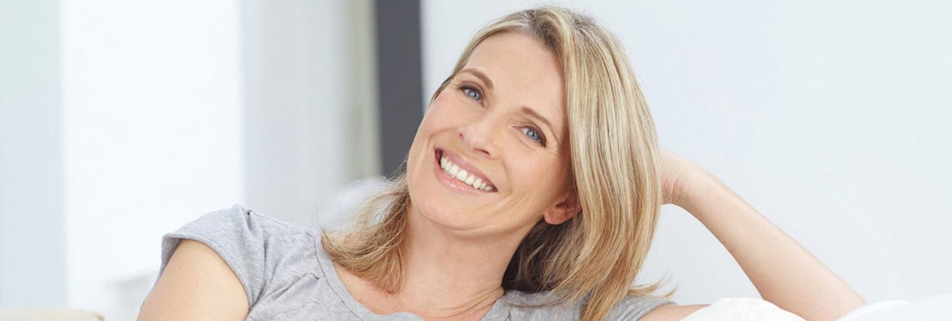 Mulher loira de meia idade com aparência jovem sorri sentada em sofá com a cabeça apoiada na mão