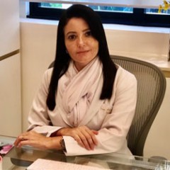de jaleco branco cabelos pretos na altura do peito sentada na mesa do consultório dra amanda nabuco olha pra câmera com expressão neutra