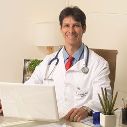 medico de cabelo preto jaleco branco e estetoscópio sorri para câmera sentado na mesa em frente ao computador